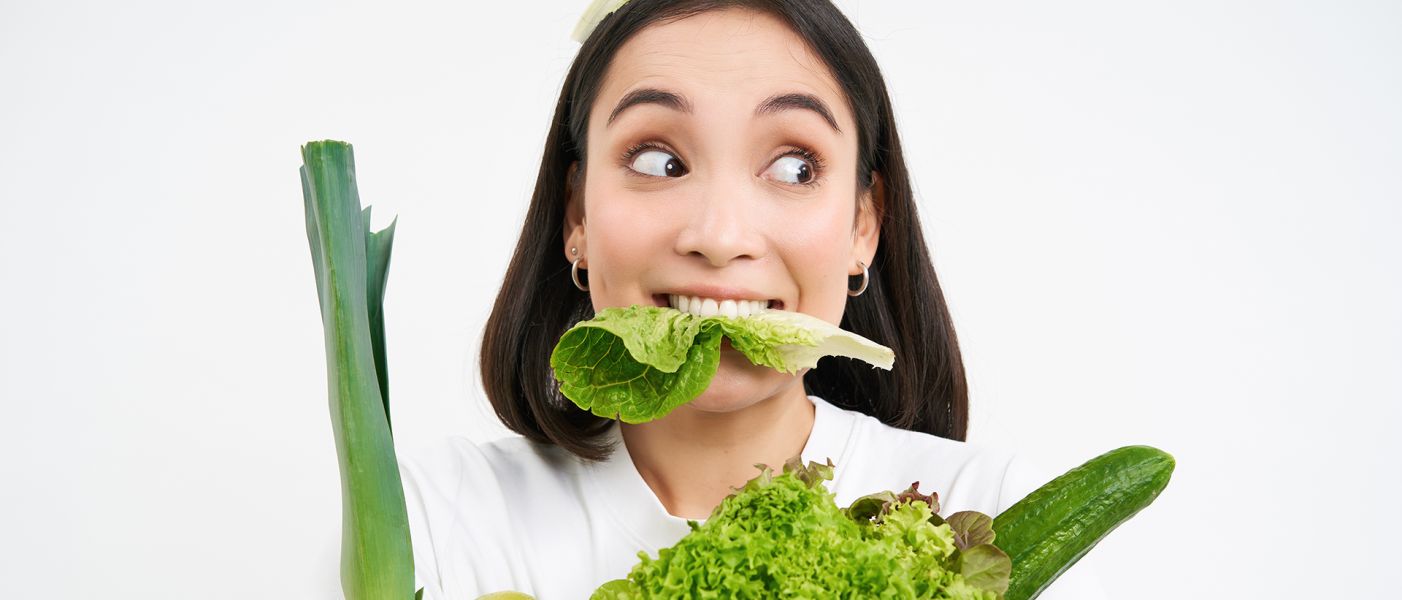 Ritratto ravvicinato di una ragazza asiatica che mastica una foglia di lattuga, con aria emozionata, tenendo in mano un mucchio di verdure, cibo biologico verde dall'orto, in piedi su sfondo bianco